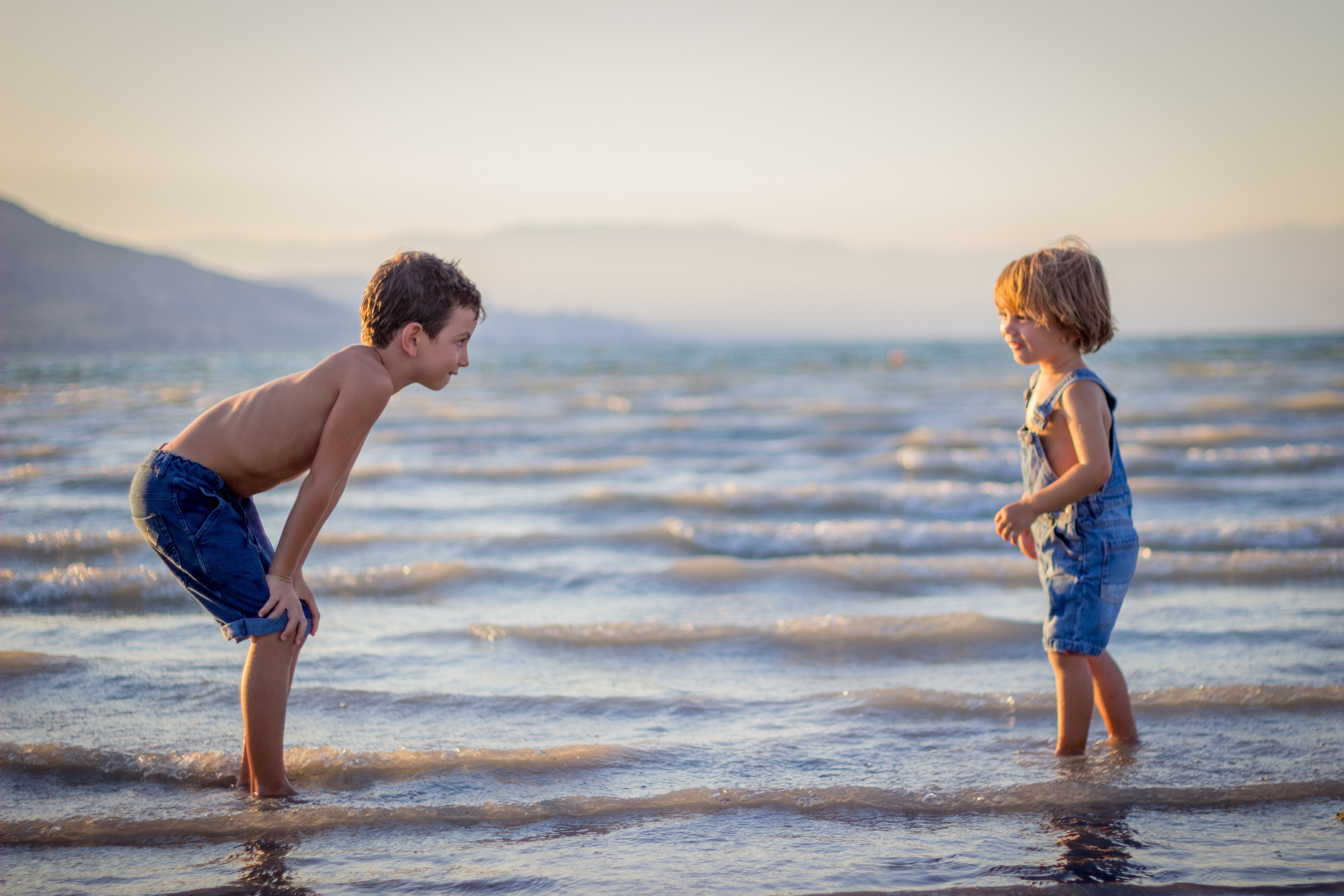       Duas crianças de pele clara, uma de frente à outra, estão com os pés na beira do mar 
