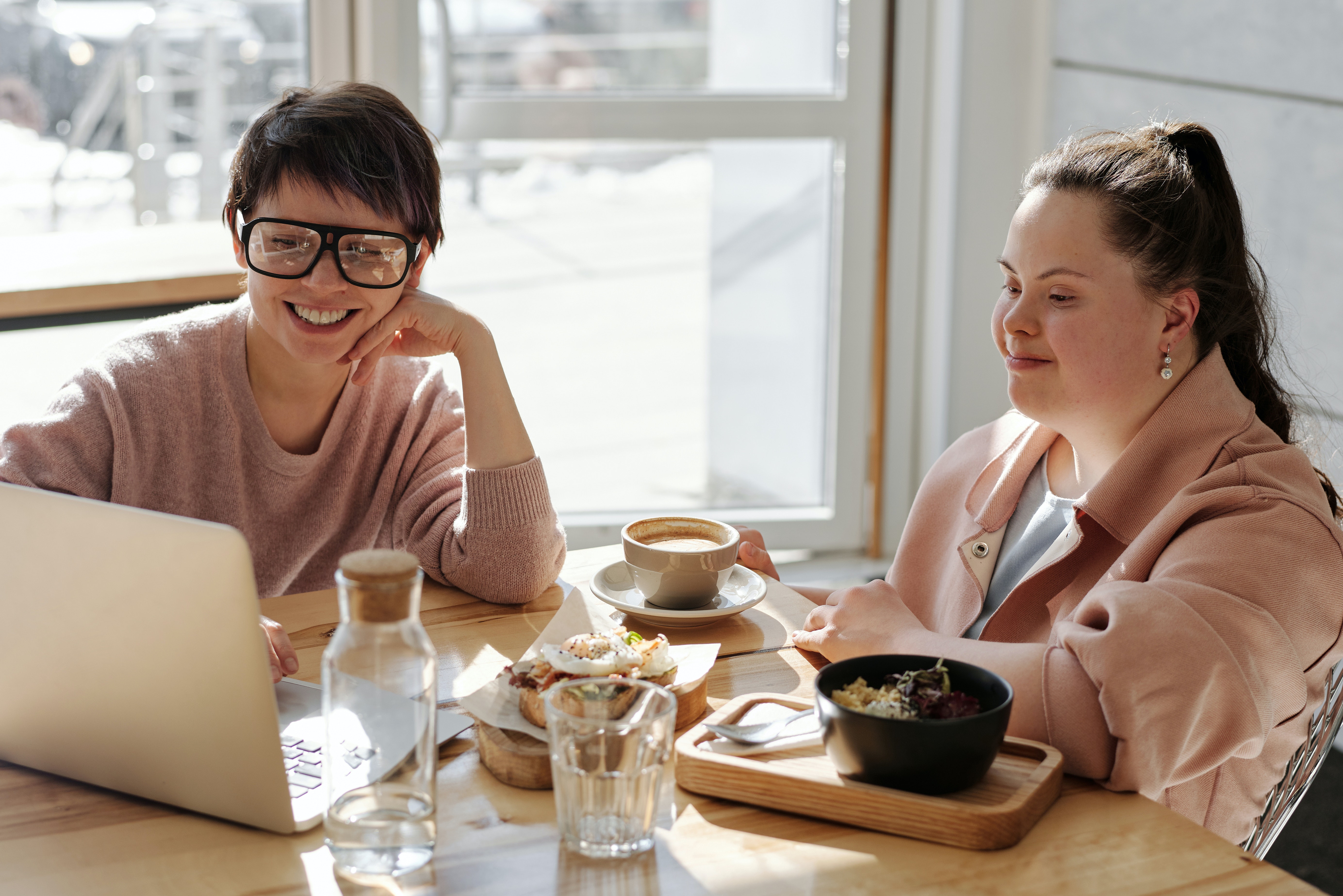 mulher branca com síndrome de down sentada à mesa com outra mulher branca, que usa óculos. Elas sorriem enquanto olham para tela do notebook.